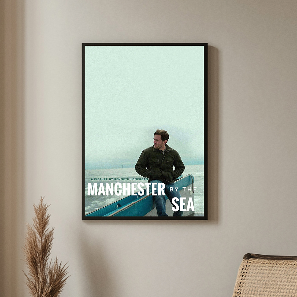 Framed poster of Manchester by the Sea featuring a man sitting thoughtfully by the sea in a minimalist design, displayed on a neutral-colored wall.