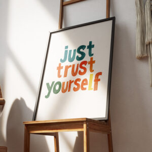 Framed poster with the motivational quote 'Just Trust Yourself' in colorful, bold letters against a white background, leaning on a textured concrete wall.